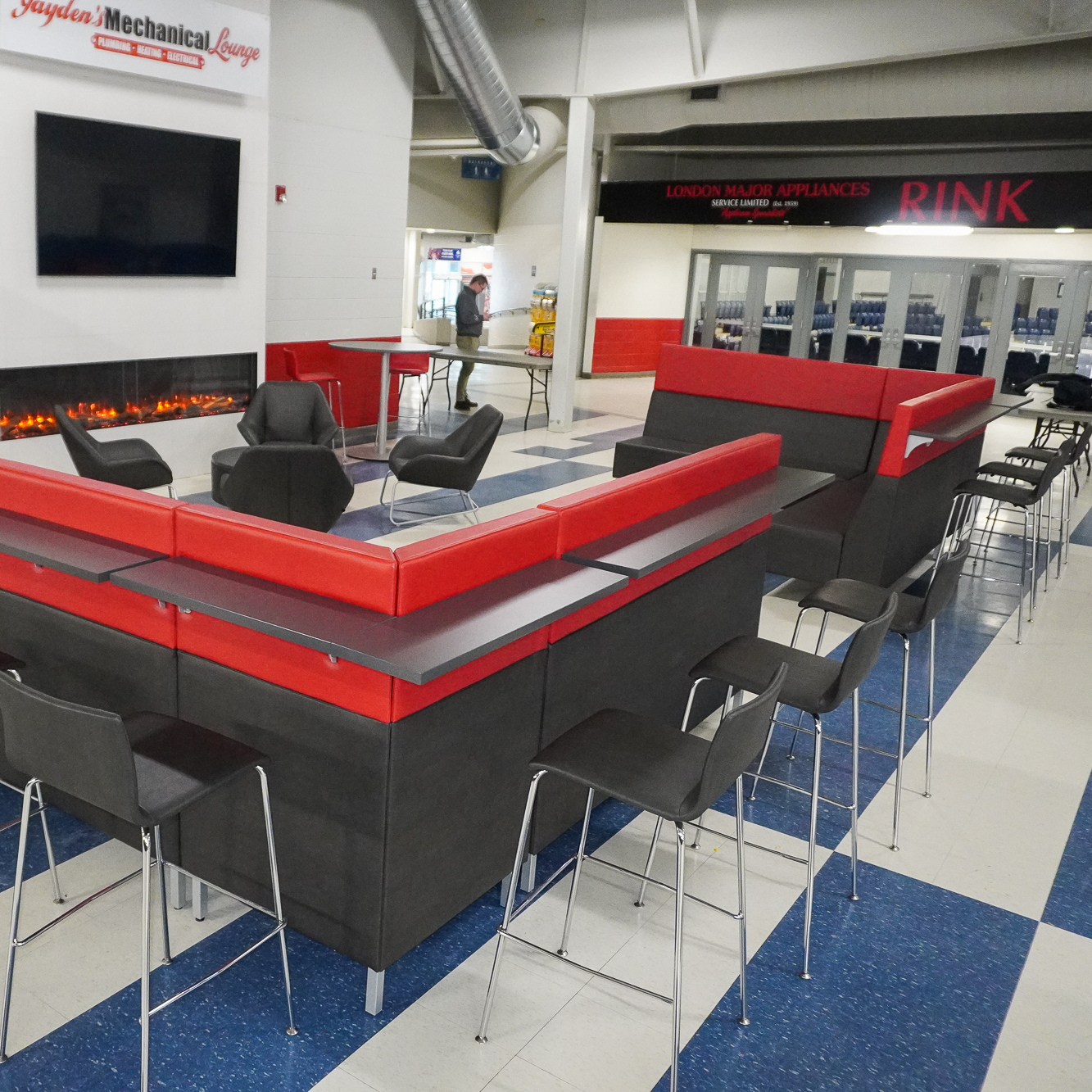 Modern arena lounge featuring custom red and black banquette seating with integrated counters, high stools, and café tables, centered around a contemporary linear fireplace with wall-mounted TV, designed for social gathering and spectator comfort.